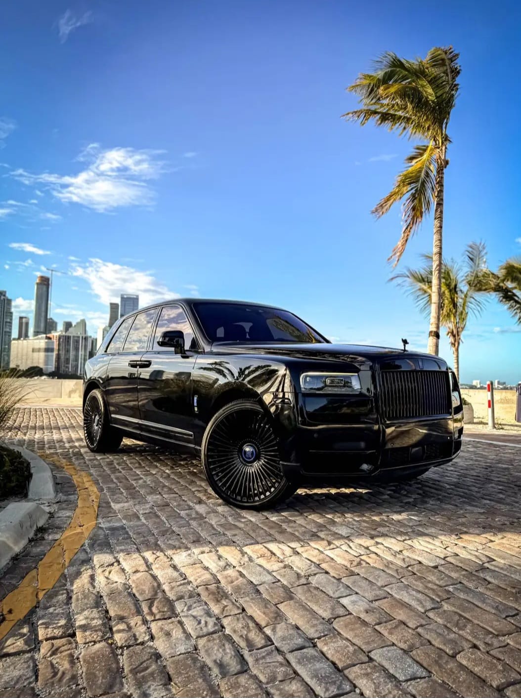 Blacked-out Rolls-Royce Cullinan parked on cobblestone with palm trees at dusk